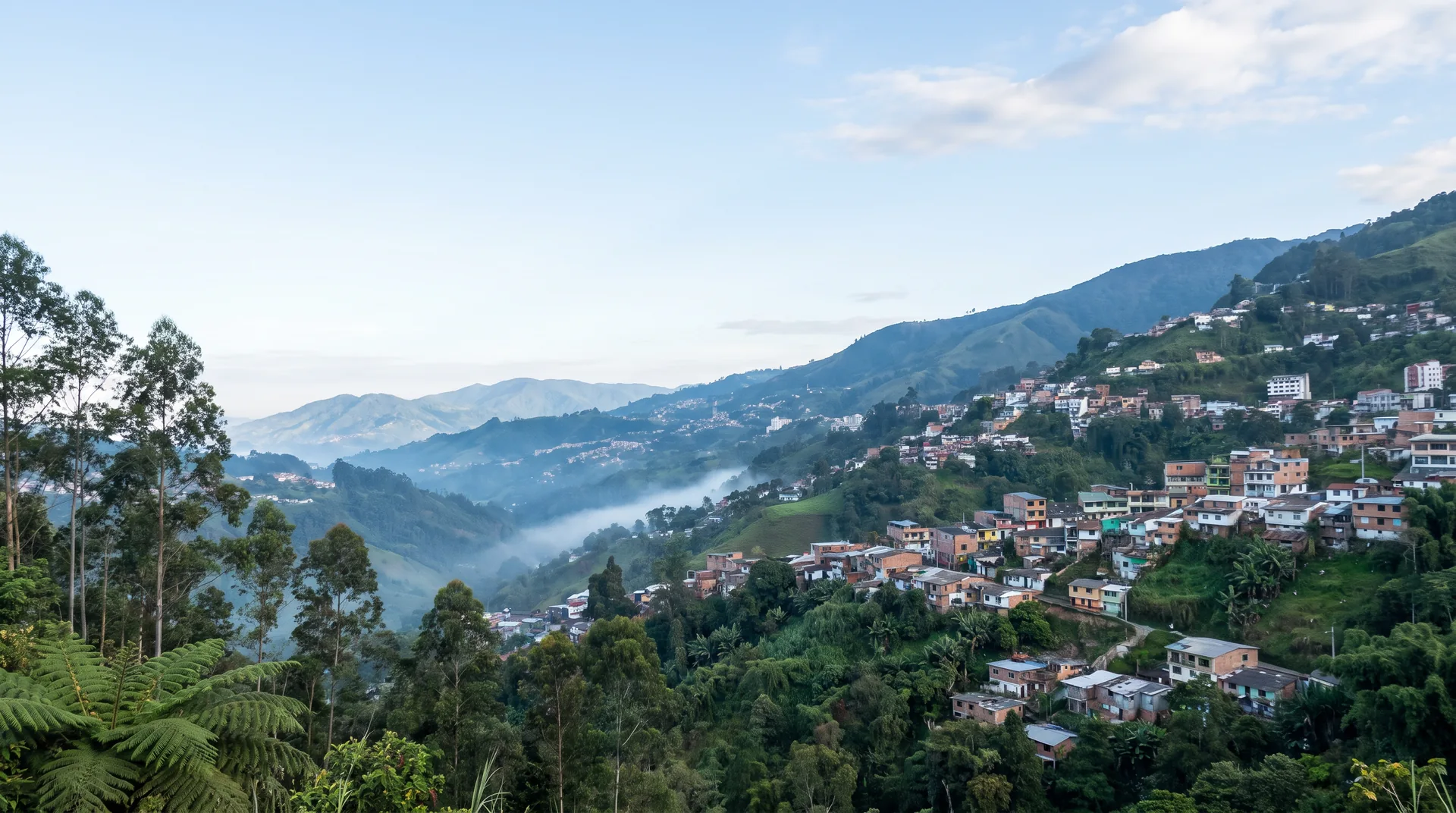 Imagen ilustrativa de una ciudad de ladera en los Andes de Caldas: casas de colores subiendo la montaña, una tira de neblina en el valle, eucaliptos y helechos en primer plano